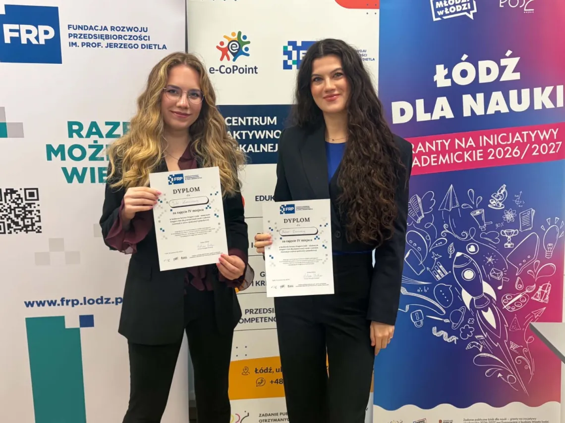 Dwie młode kobiety w ciemnych garniturach trzymają dyplom, z tyłu rolllupy organizatorów