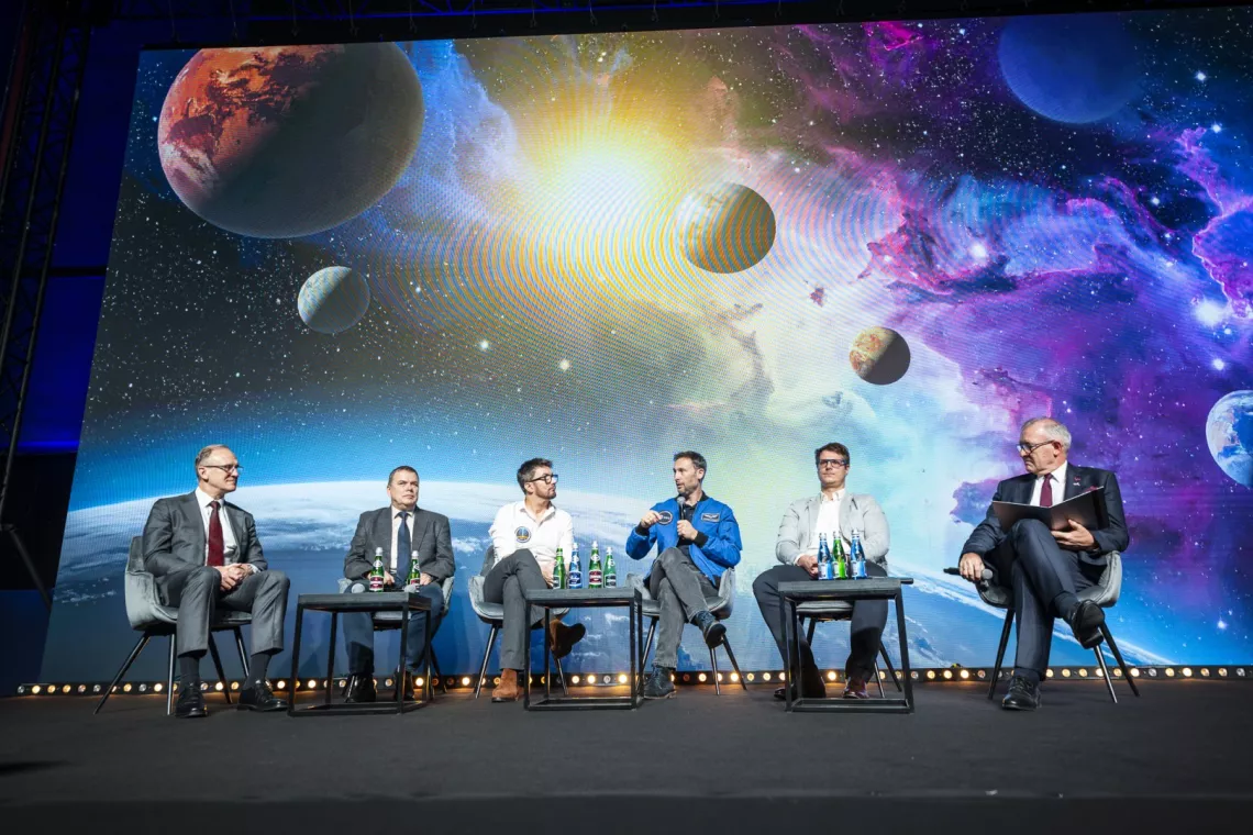 Panel ekspercki, w tle ekran z wizualizacją planet w kosmosie