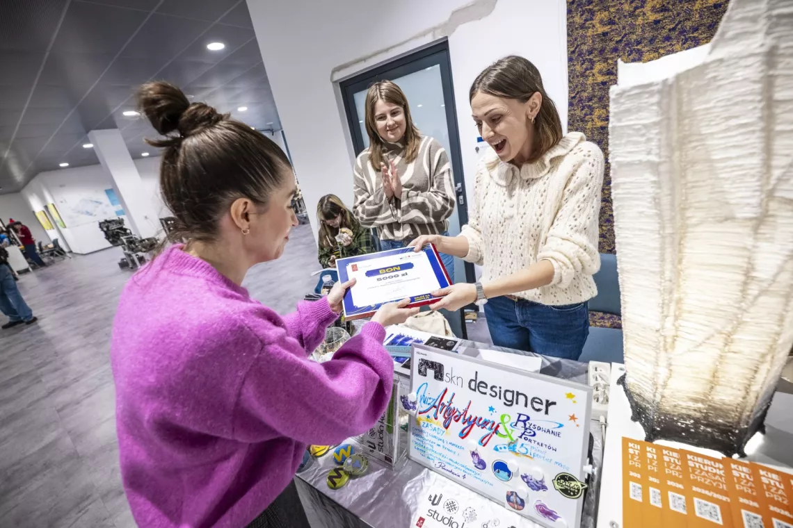 Kobieta w fukssjowym swetrze wręcza czek z nagrodą, który odbiera uradowana osoba