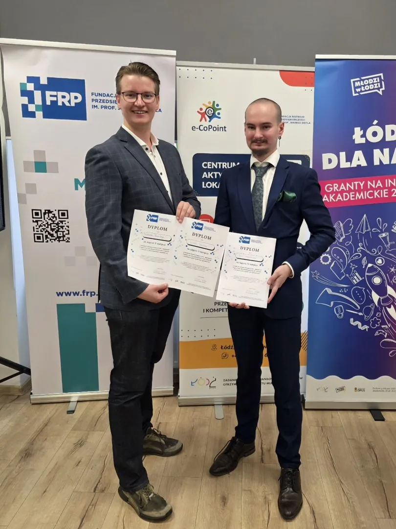 Dwóch młodych mężczyzn w garniturach z dyplomami w ręku, z tyłu rollupy organizatorów