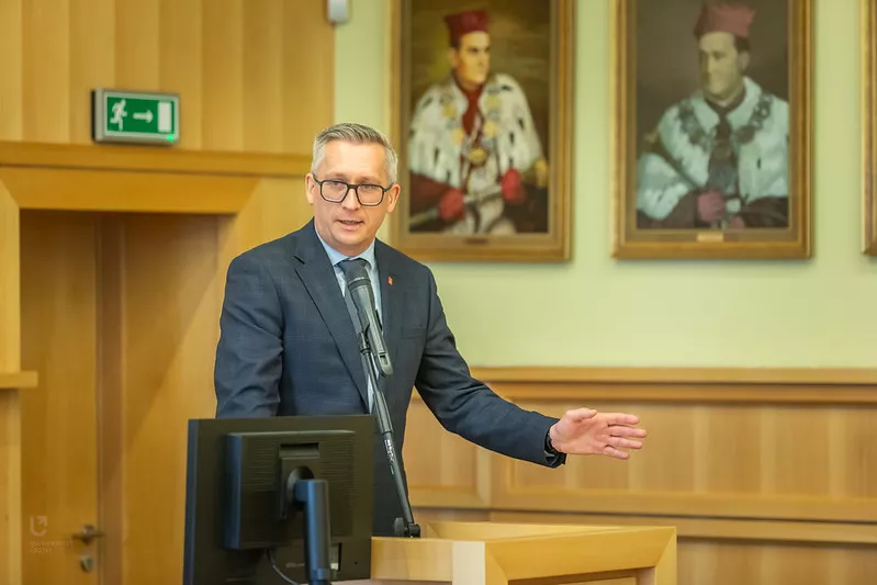 Elegancko ubrany mężczyzna w okularach przemawia przy mównicy, w tle widoczne obrazy rektorów w uroczystych strojach