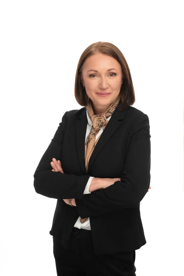 Kobieta z włosami do ramion, w ciemnym garniturze, apaszce na szyi, z rękoma skrzyżowanymi w ramionach