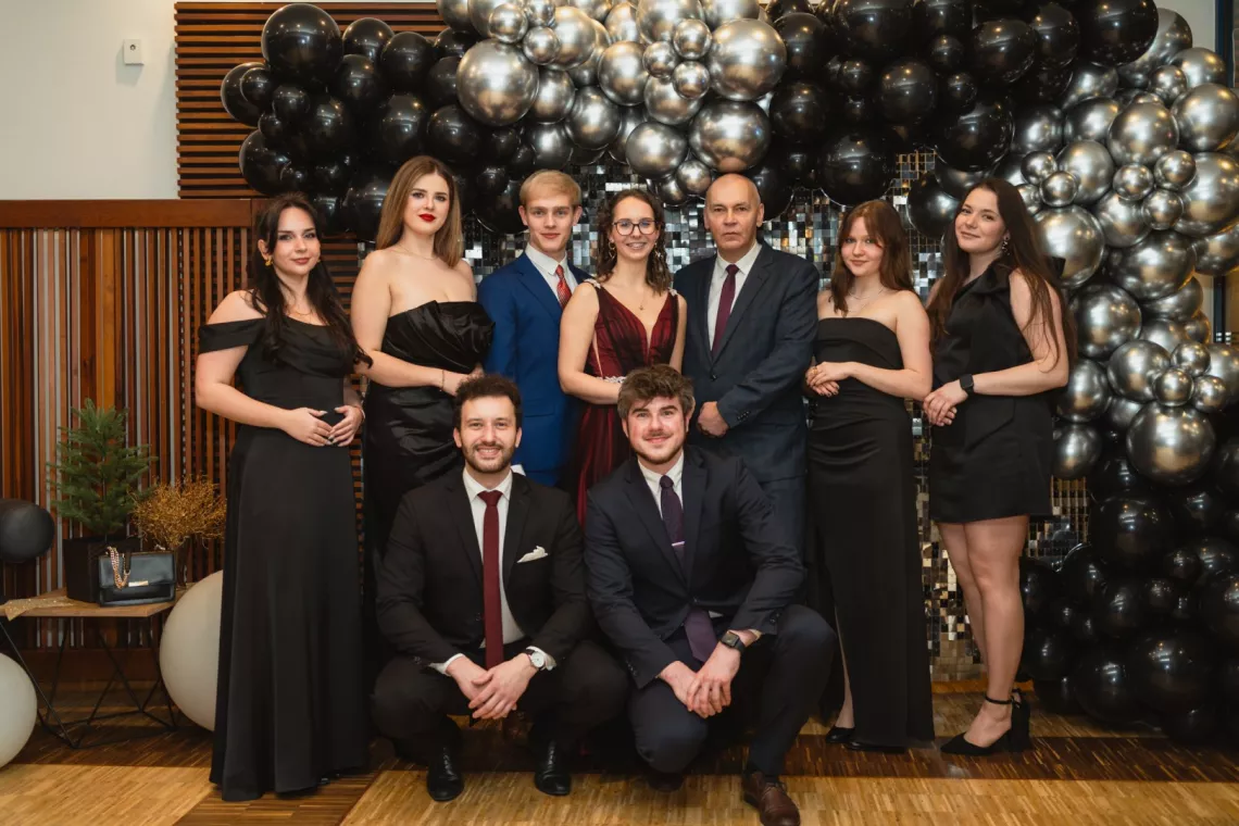 Wieczorow ubrane osoby stoją na tle ścianki ułożonej z czarnych i srebrnych balonów