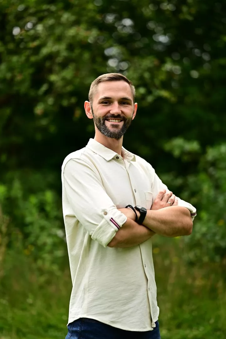 Mężczyzna z brodą w jasnej koszuli trzyma ręce skrzyżowane na piersiach, wokół dużo zieleni