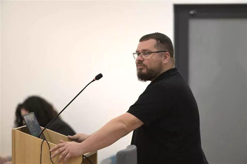 Mężczyzna z brodą i w okularach wypowiada się do mikrofonu stojącego na mównicy. Ubrany jest w czarny tshirt.