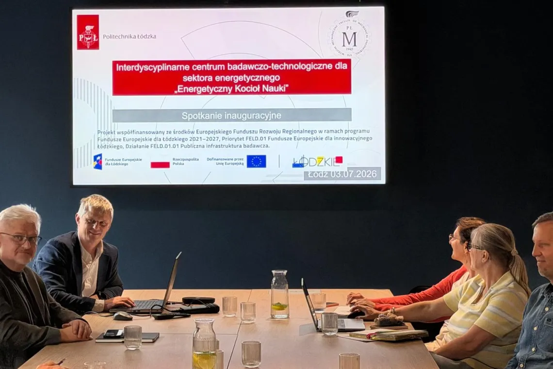 Grupa osób przy stole konferencyjnym, na którym znajdują się laptopy i szklanki z napojami, w tle na czarnej ścianie ekran z nazwą projektu i logo instytucji dofinansowujących oraz beneficjentów