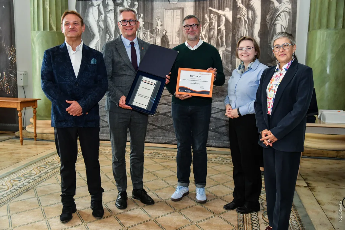 Dwie kobiety i trzech mężczyzn pozuje do zdjęcia w historycznej sali z kolumnami i rzeźbami, dwóch mężczyzn trzyma dyplomy