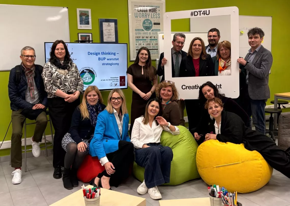 W pomieszczeniu z pufami i zielonymi scianami kilkanaście uśmiechniętych osób pozuje do zdjęcia