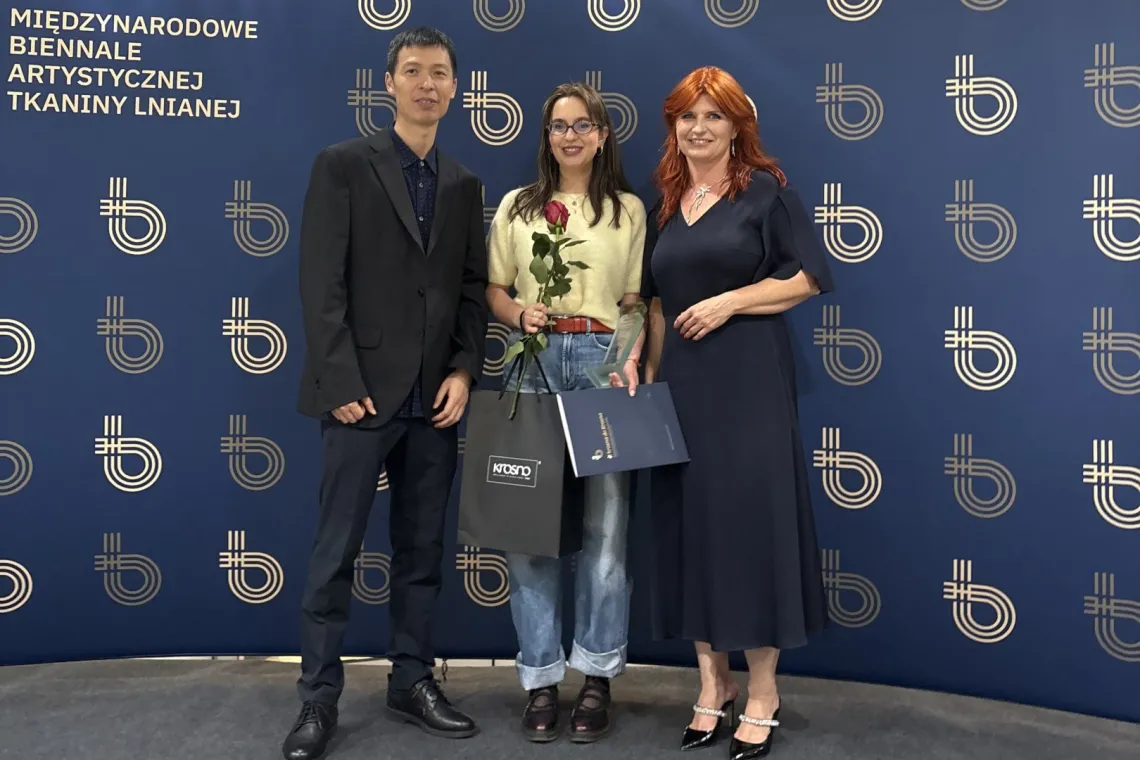 Troje uczestników wydarzenia pozuje do zdjęcia na tle ścianki z logo Międzynarodowego Biennale Artystycznej Tkaniny Lnianej; jedna z osób trzyma różę i pamiątkowy dyplom.