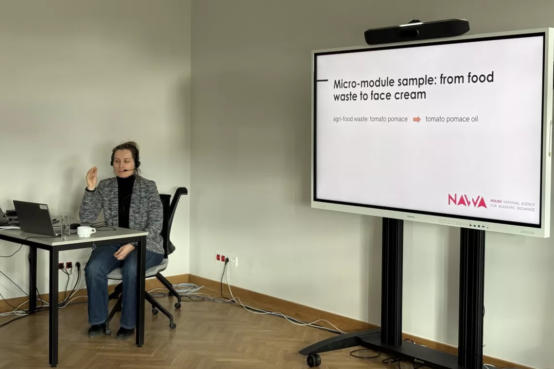 Młoda kobieta siedzi za biurkiem wykładowcy i prowadzi wykład, obok na ekranie wyświetla się temat wystapienia