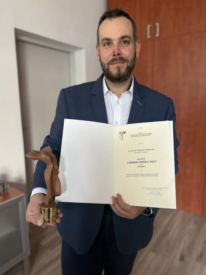 Mężczyzna w garniturze i białej koszuli trzyma statuetkę oraz dyplom, stoi w gabinecie 