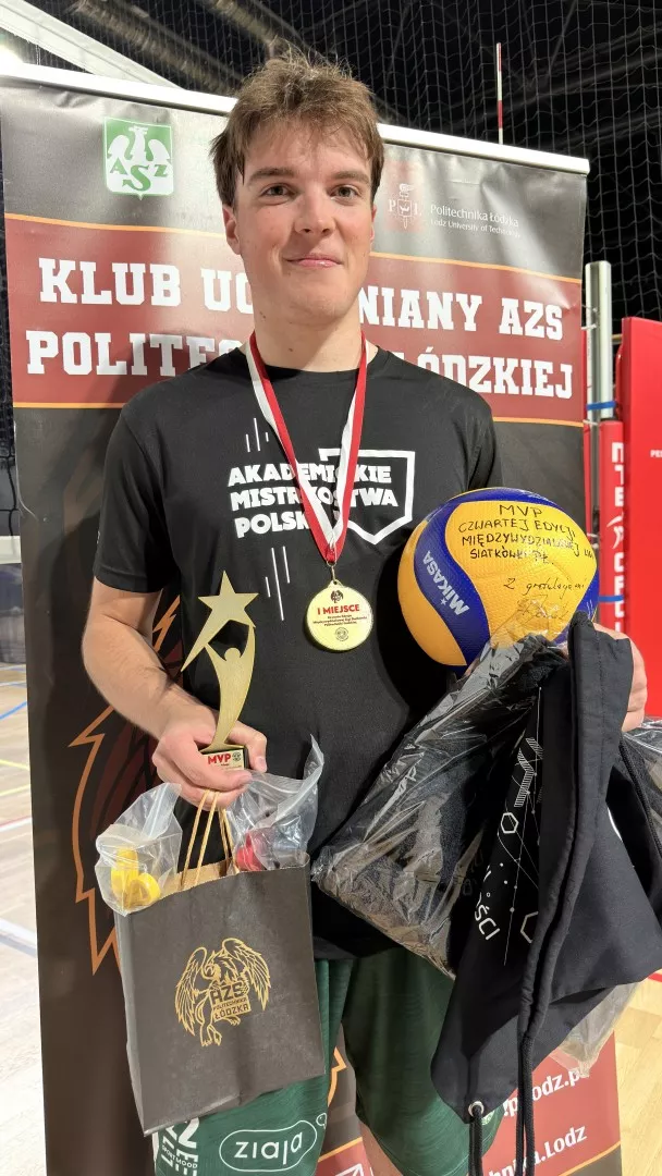 Młody chłopak z czarnej koszulce z medalem na piersi, piłką siatkarską w ręku oraz nagrodami