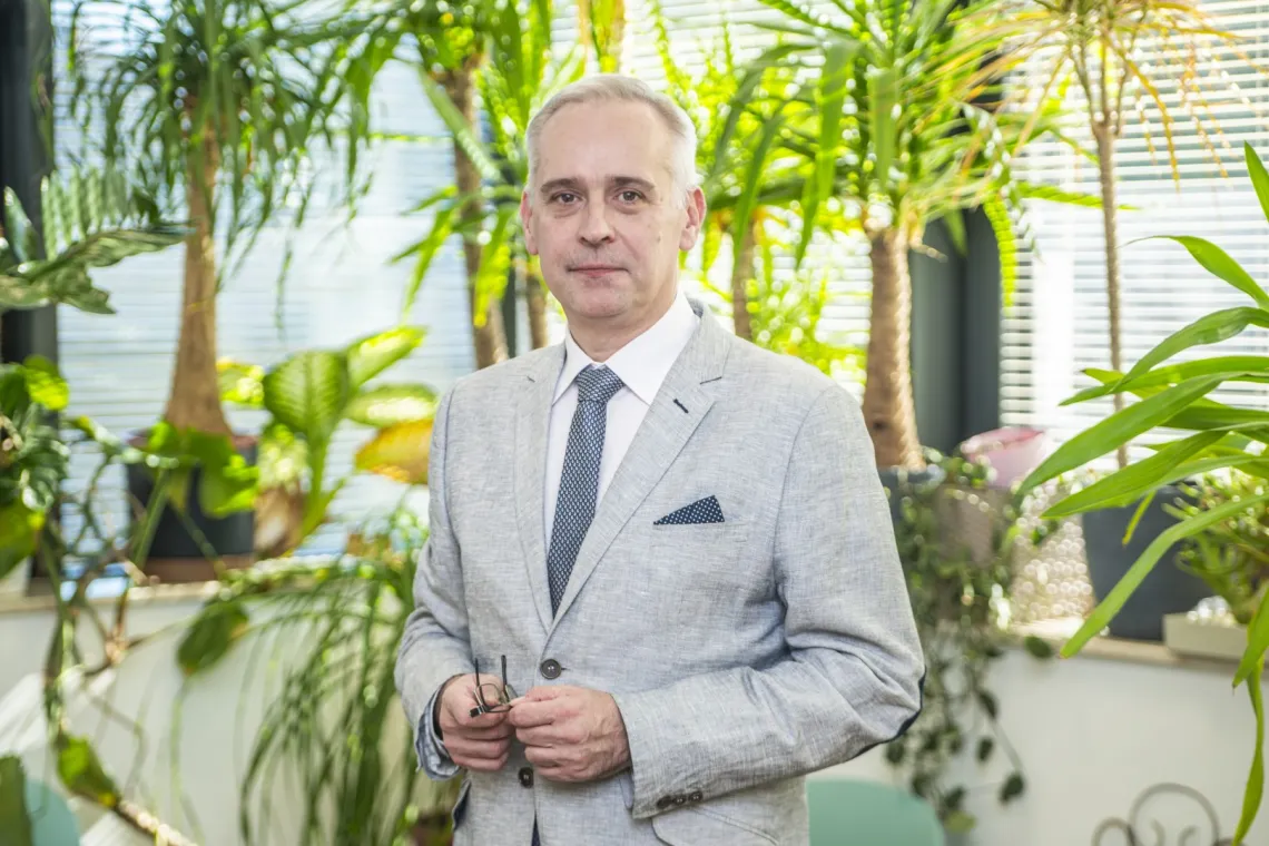 Elegancko ubrany mężczyzna, szara marynarka, biała koszula, ciemny krawat i poszetka,  trzyma w ręku okulary, w tle duże rośliny doniczkowe stojące na oknie