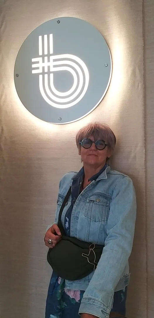 Kobieta w okularach, ubrana w dzinsowej kurtce i kolorowej sukience, z torba przewieszona przez ramię stoi pod oświetlonym logo