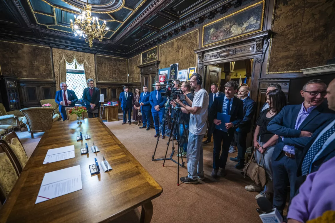 Liczna grupa oób w historycznym wnętrzu, widać także dwóch operatorów kamer. Stoją, czekając na rozpoczęcie spotkania