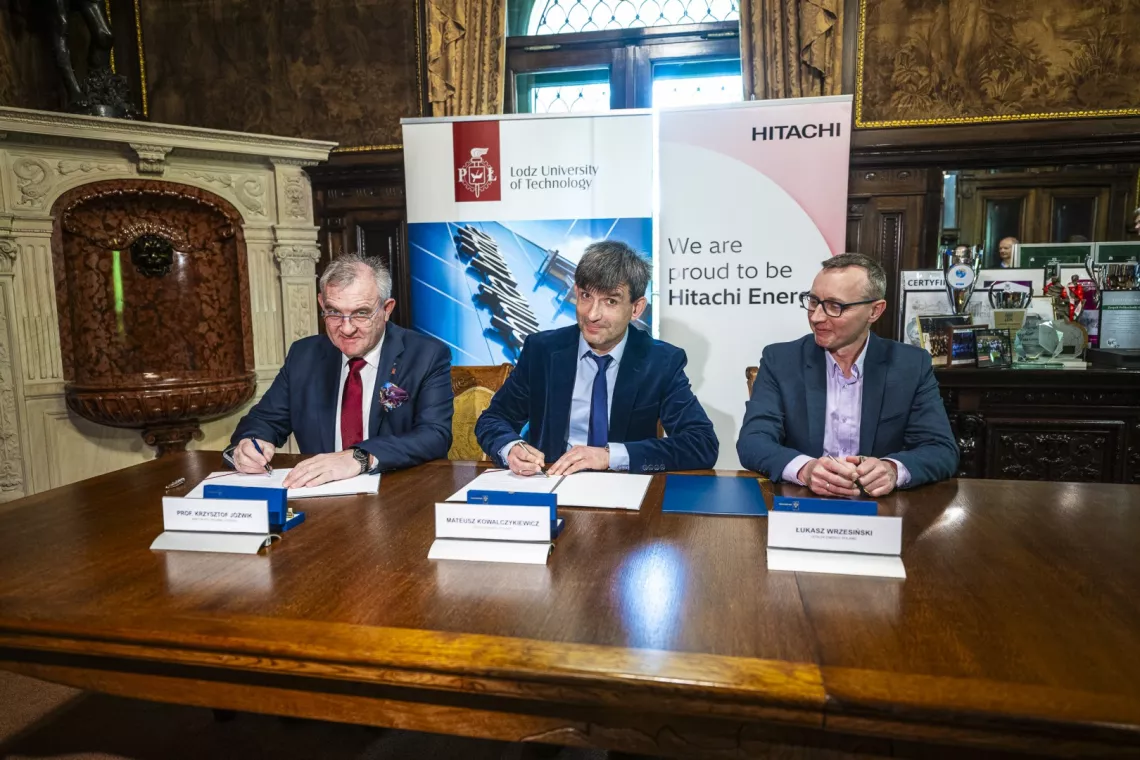 Trzech mężczyzn siedzi przy stole, na którym leżą dokumnty do podpisu. Z tyłu rollupy z napisem Politechnika Łódzka i Hitachi