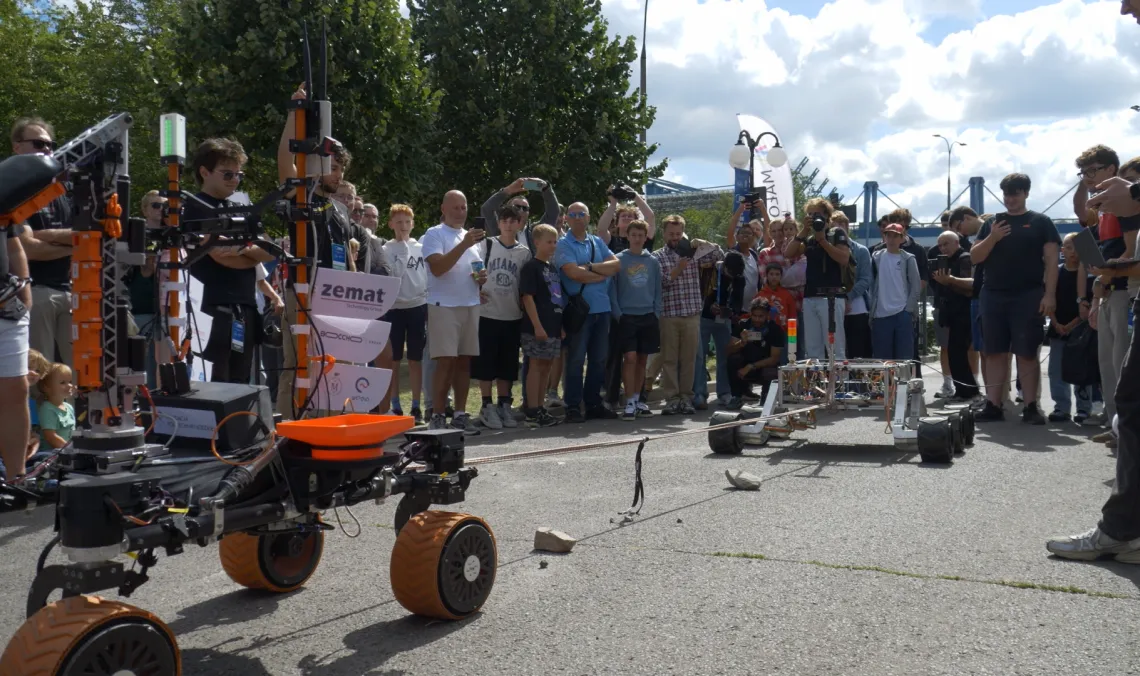Dwa roboty mobilne prezentowane podczas pokazu przed zgromadzoną publicznością; w tle widoczna zieleń i pochmurne niebo
