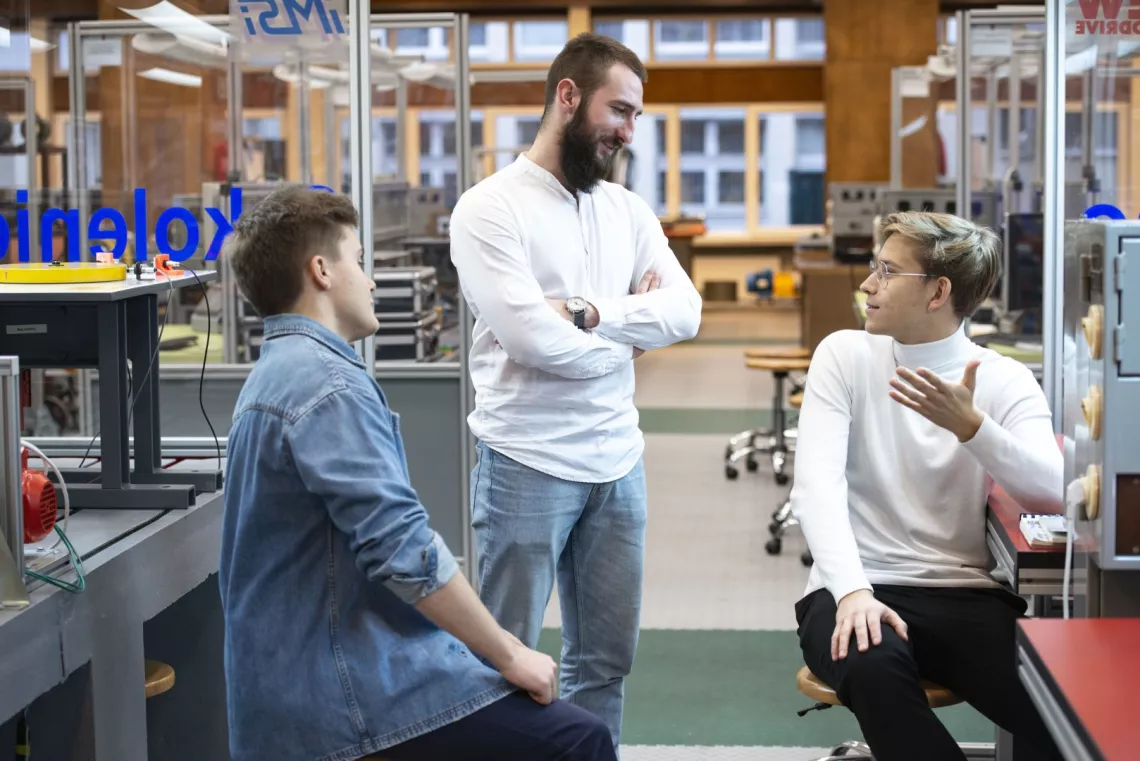 W laboratorium  machatronicznym rozmawia trzech studentów, siedzących przy stanowiskach