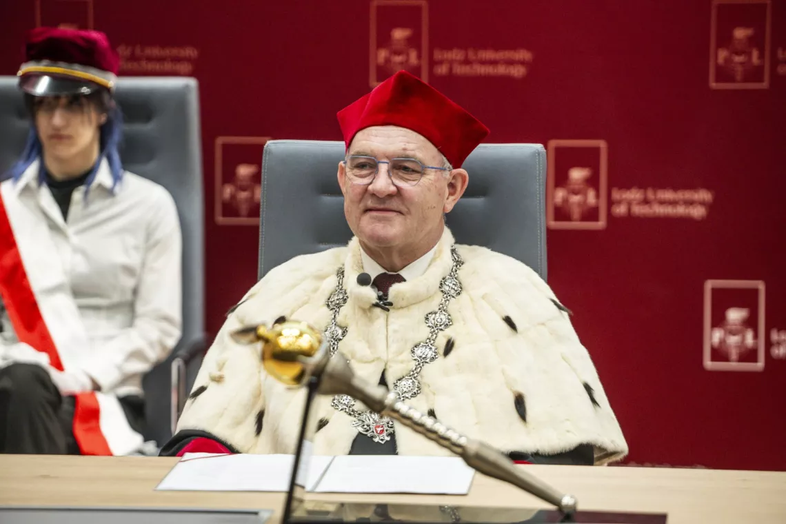 Rektor siedzi za katedrą w uroczystym stroju, przed nim widać godło rektorskie
