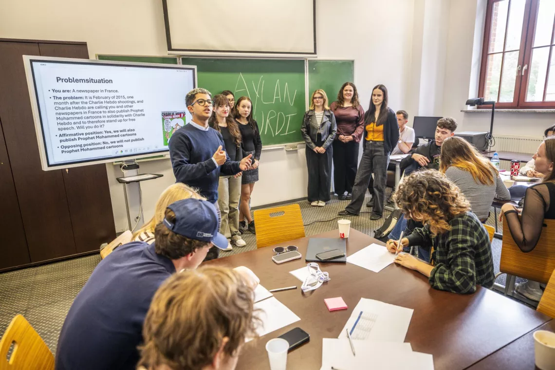 Debata prowadzone w ramach programu przrez moderatora stojącego przy ekranie. Wokół grupa studentów przy tablicy i przy stołach