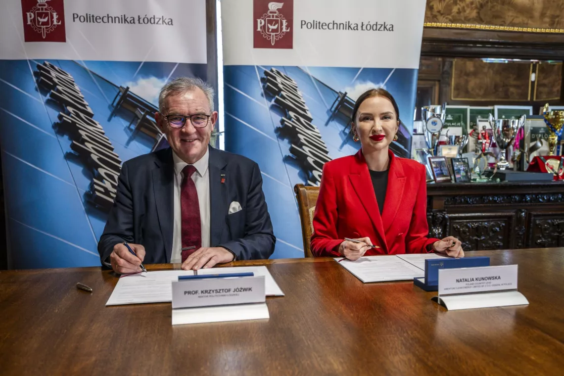 Kobieta w czerwonej marynarce i mężczyzna w garniturze patrzą w obiektyw, przed nimi leżą dokumenty do podpisania