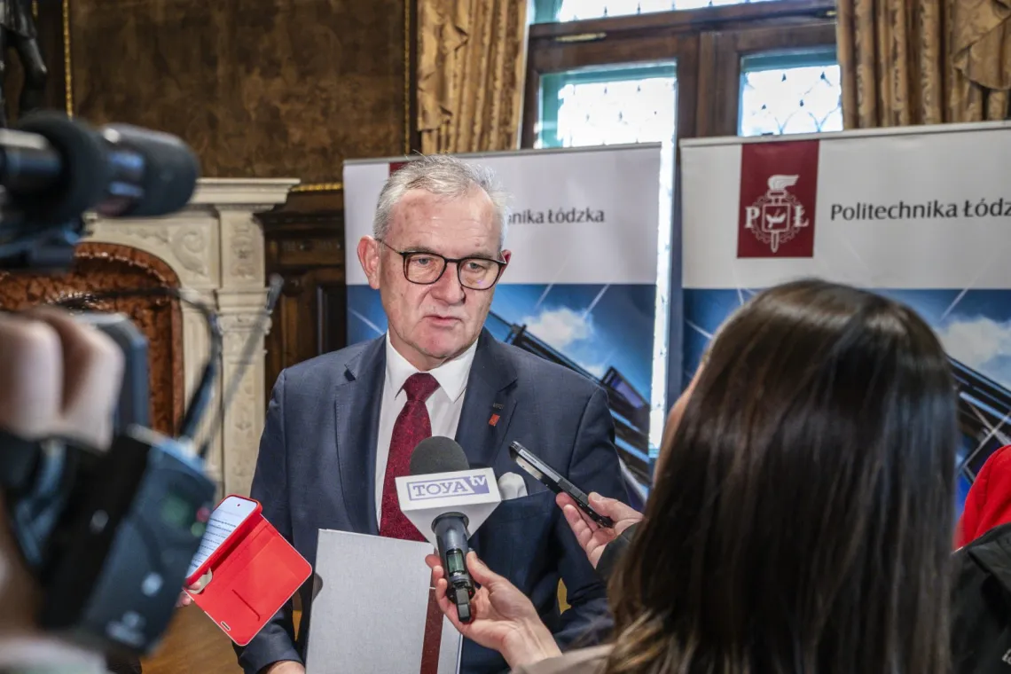 Mężczyzna w garniturze, w okularach udziela wywiadu dziennikarzom radia i telewizji. Zabytkowe wnętrze, w którym ustawione są rollupy z napisem Politechnika Łódxka