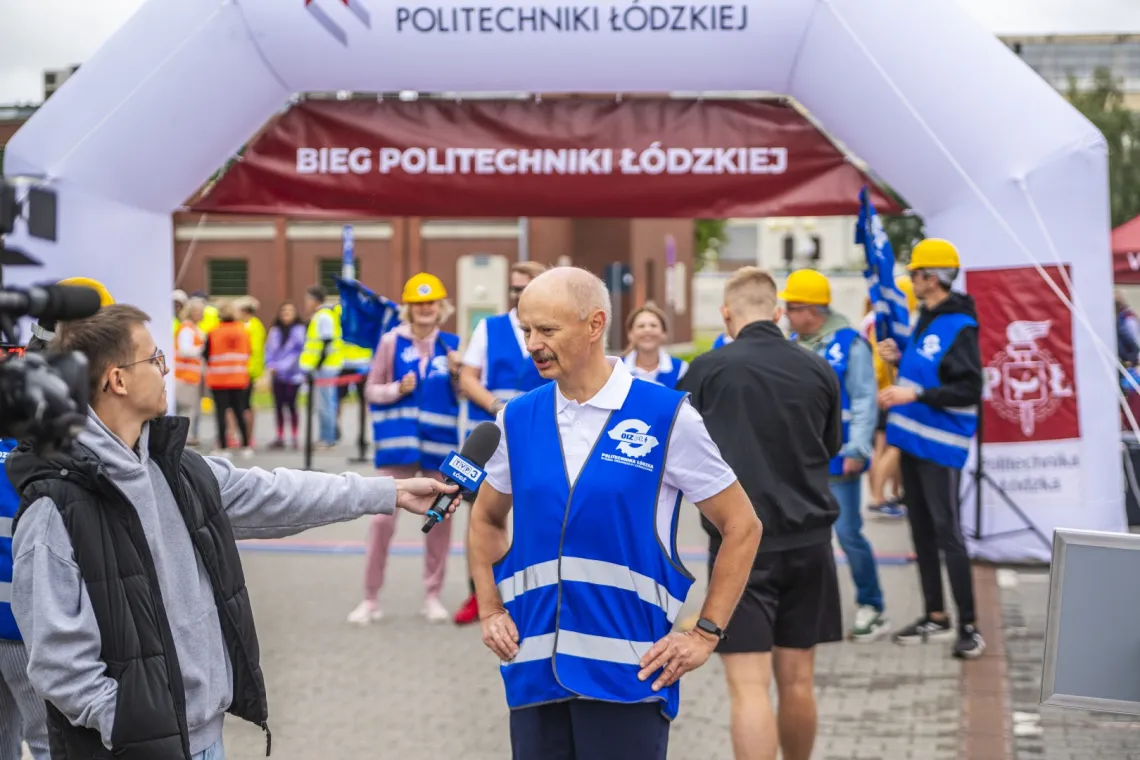 Mężczyzna w niebieskiej kamizelce w białe paski z napisem OIZ rozmawia z dziennikarzem trzymającym mikrofon z napisem TVP3. W tle uczestnicy biegi i dmuchaniec z napisem Bieg Politechniki Łódzkiej