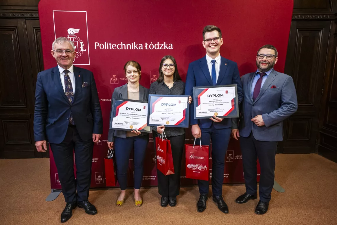 Pięć osób, trzy z nich trzymają dyplomy, stoi na tle banera z napisem Politechnika Łódzka 