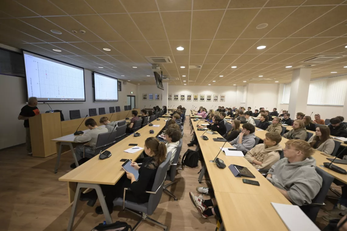 Młodzież siedzi w Sali Senatu, słuchając nauczyciela stojącego przy mównicy