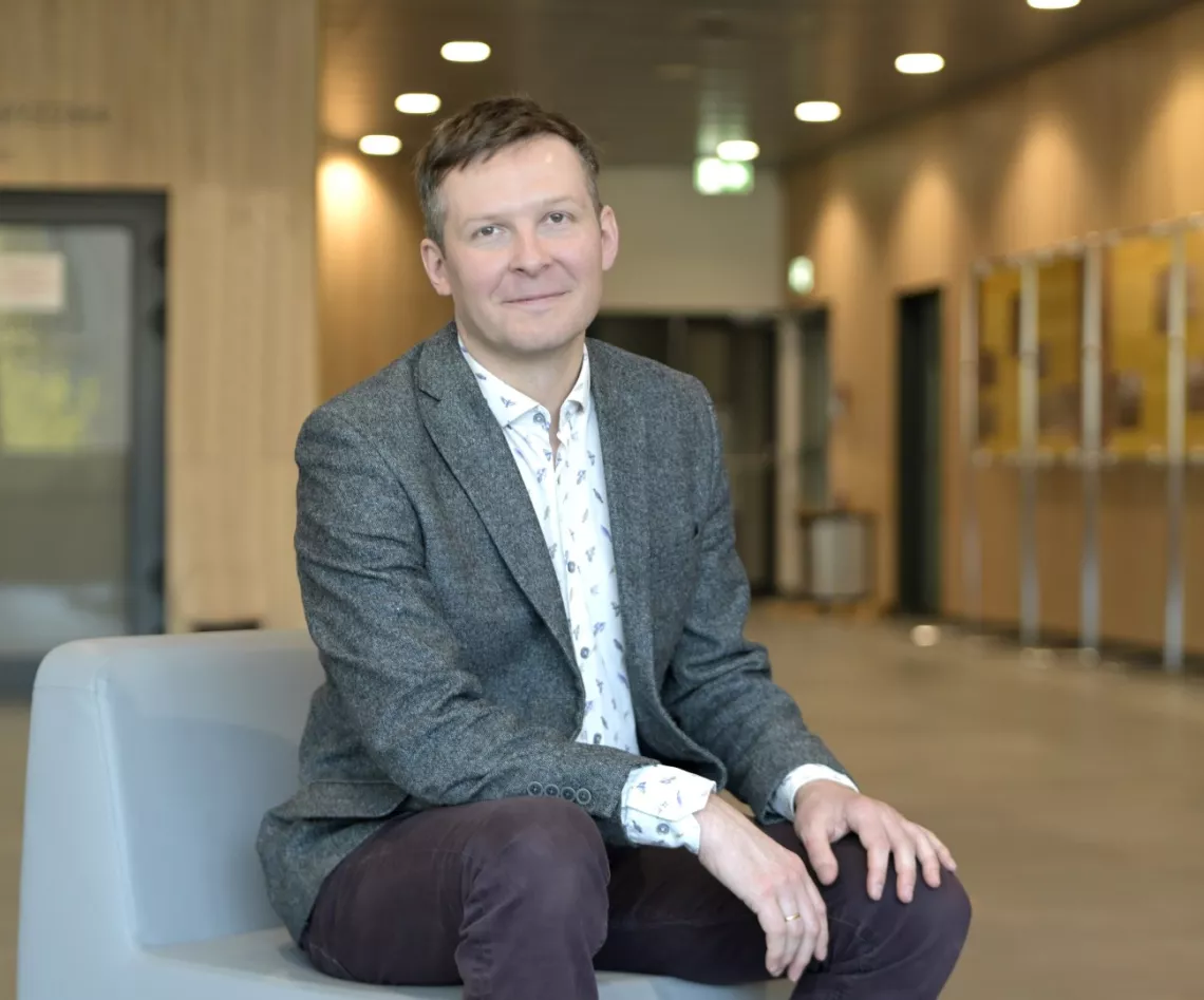 Młody mężczyzna w szarej marynarce siedzi w holu budynku, trzymając ręce na kolanach