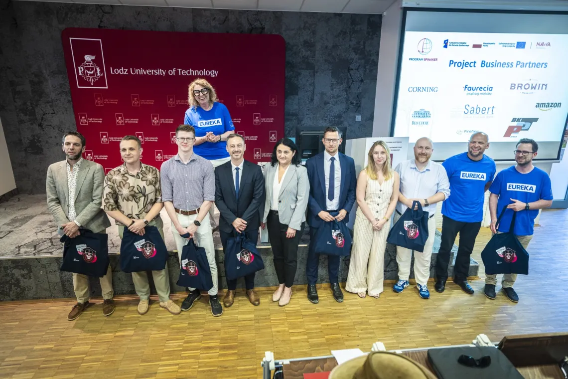 Grupa 10 osób stoi przed podium, trzymając w rekach materioławe torby, prawdopodobnie z prezentami. Za tą grupą jest usmiechnięta kobieta w niebieskiej podkoszulce z napisem EUREKA oraz bordowa ścianka promocyjna z napisem Politechnika Łódzka