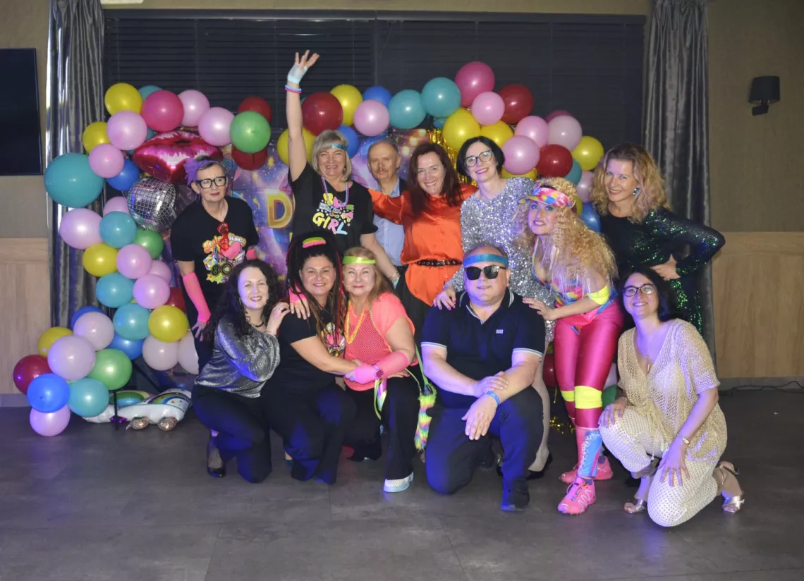 Grupa roześmianych osób, w ubraniach w stylu dyskoteki z lat 80. W tle girlanada z balonów.