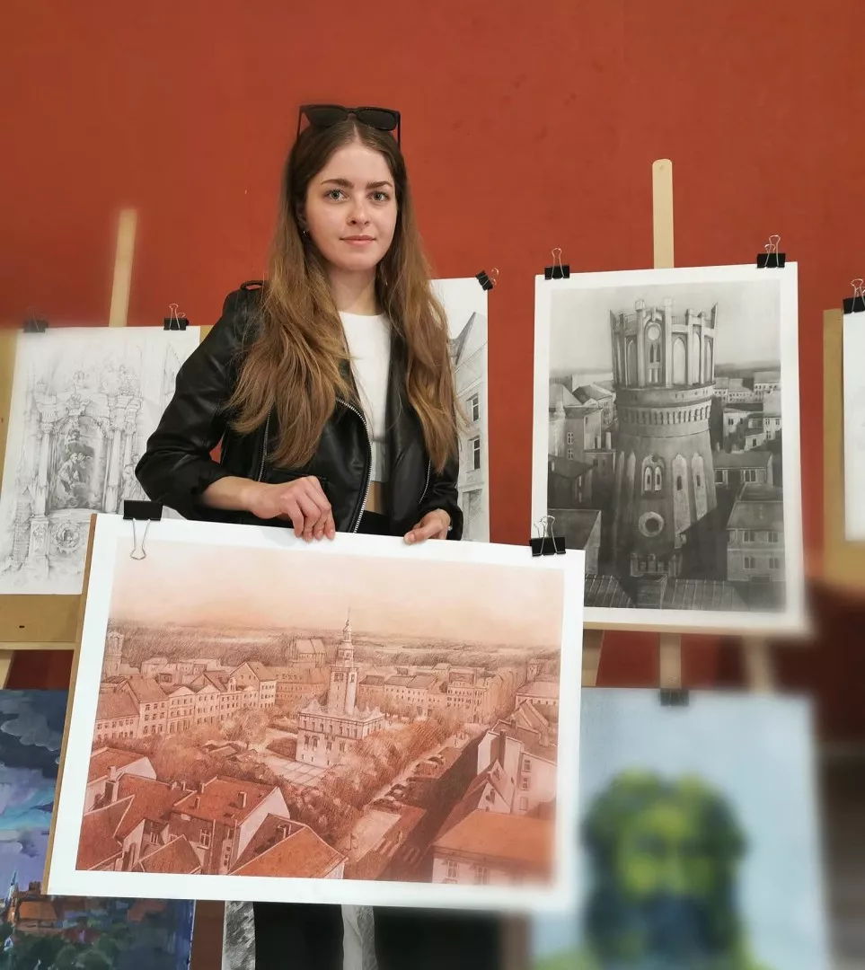 Młoda dziewczyna z długimi włosami trzyma w ręku rysunek panoramy miasta, w tle stoją na sztalugach inne prace plastyczne