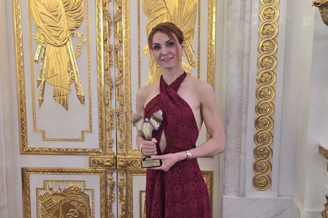 Kobieta w sukni wieczorowej trzyma w ręku statuetkę. W tle bogato złocone drzwi pałacowe. 