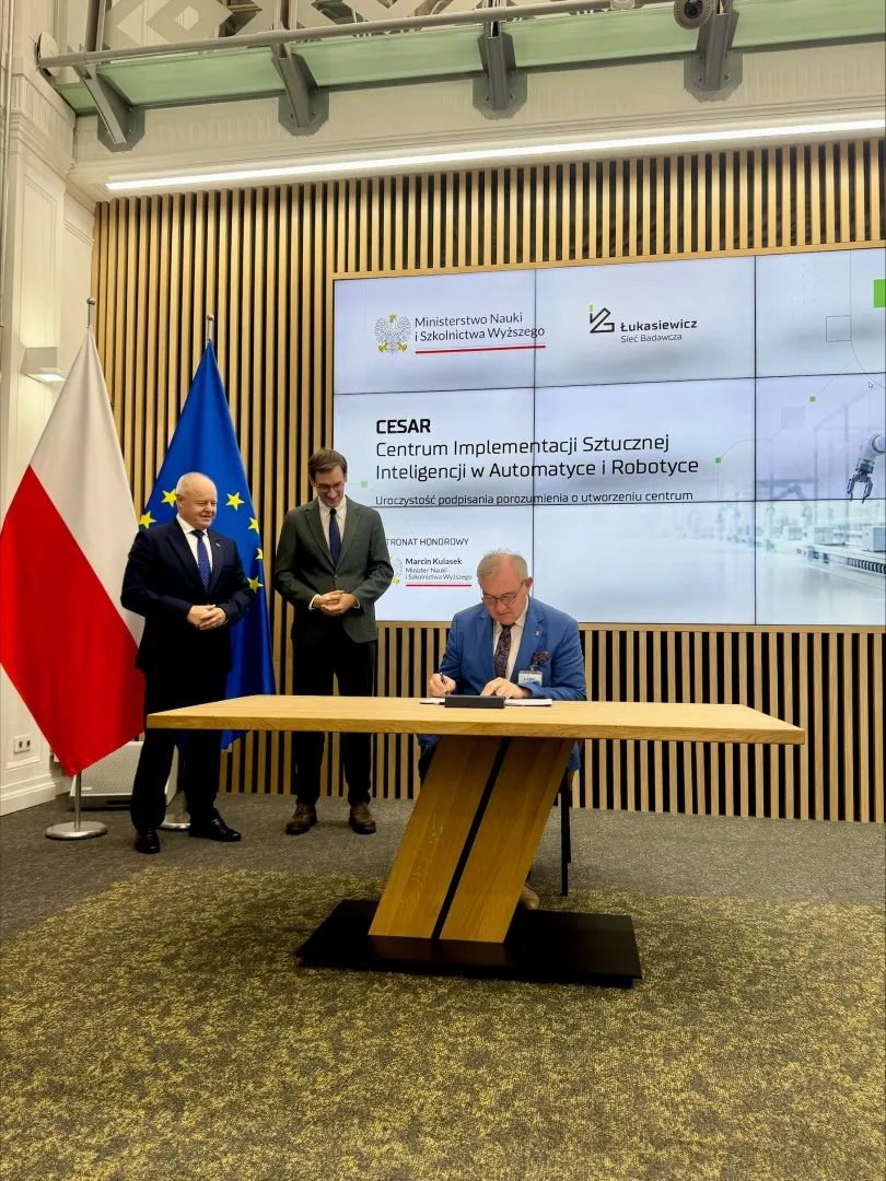 Mężczyzna w niebieskim garniturze i okularach składa podpis na dokumenci. Obo stoją dwaj  męzczyxnie w ciemnych garniturach, za którymi widać flaę Polski i UE. Z tyłu na duzym ekranie wyświetal się tytuł zawartego porozumienia.