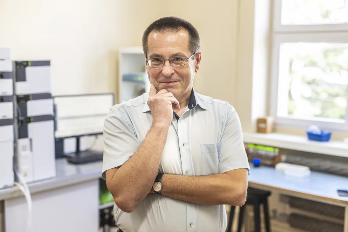 Mężczyzna w jasnej koszuli podpiera dłonią twarz, stojąc na tle nowoczesnego laboratorium 