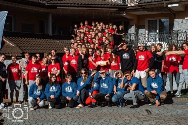Liczna grupa młodych osób pozuje do zbiorowego zdjęcia. Większość ma czerwone bluzy. W pierwszym rzędzie grupa osób w przyklęku ma na sobie granatowe bluzy. W tle budynek, słoneczne oświetlenie