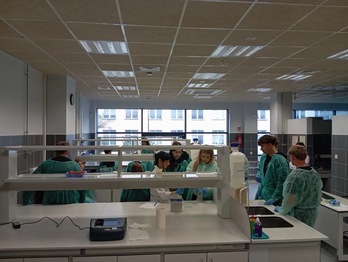 Młodziew zielonych fartuchach stoi wokól stanowiska laboratoryjnego przy którym jest wykładowca w białym fartuchu
