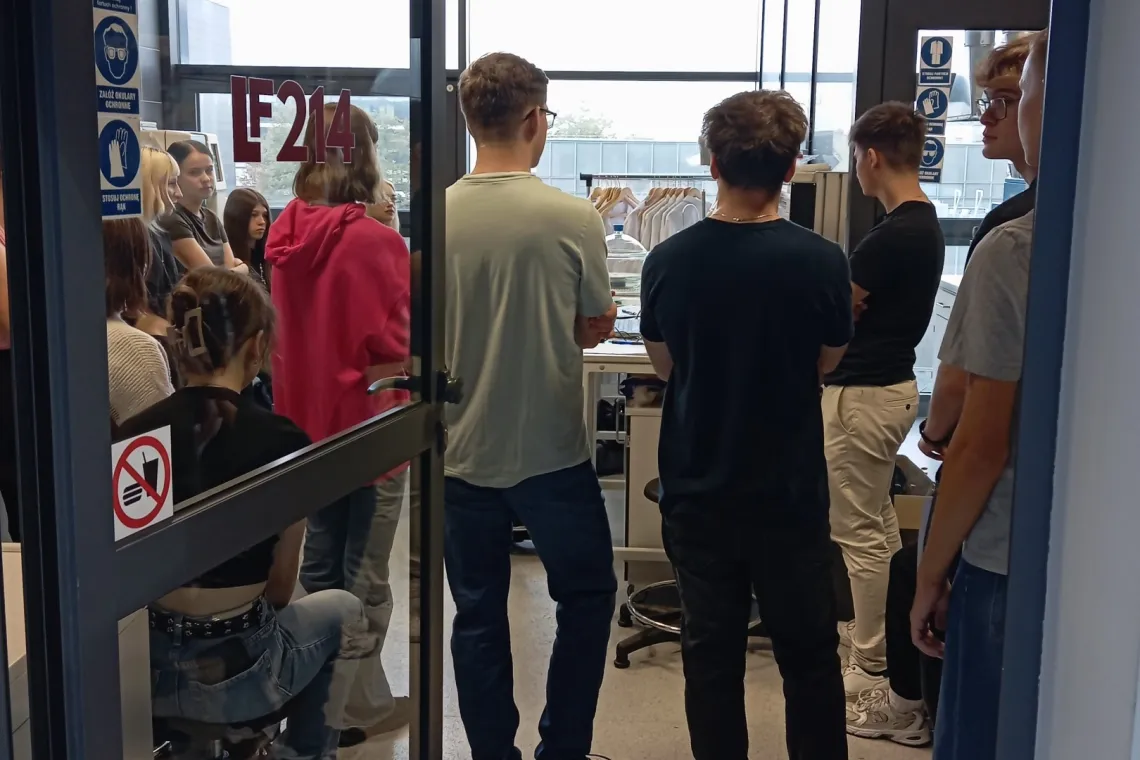 Młodzież za otwartymi drzwiami do laboratorium fizycznego słucha i przyglada się czemuś, czego nie widać na zdjęciu