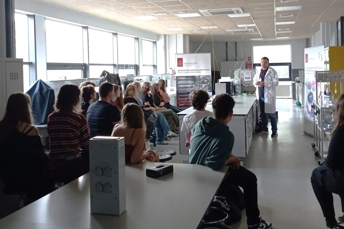Grupa młodzieży siedzi w laboratorium i słucha wykładowcy ubranego w biały fartuch