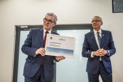 Dwóch elegancko ubranych mężczyzn stoi, jeden z nich trzyma w ręku dyplom
