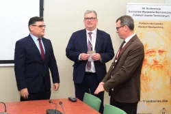 Trzech mężczyzn w garniturach rozmawia, stojąc przy stole, za nimi plakat konferencyjny 