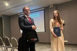 Dr inż. Magdalena Gajek (po prawej) stoi w sali konferencyjnej, trzymając w rękach granatową teczkę z logo Polskiej Akademii Nauk. Obok niej, po lewej stronie, stoi mężczyzna w garniturze. W tle widoczny ekran z napisem „Sympozjum Analityczne Gdynia 2025”.