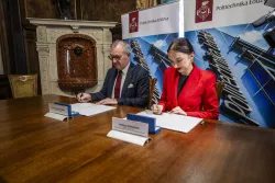 Kobieta w czerwonej marynarce i męzczyzna w szaryjm garniturze podpisuja umowę pzry stole, na którym stojąwizytówki. Z tyłu rollup z napisem Politechnika Łódzka 