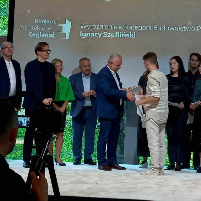 Na pierwszym planie mężczyzna w jasnym ubraniu odbiera dyplom od elegancko ubranego mężczyzny w garniturze. W tle stoi kilkanaście osób – kobiety i mężczyźni. W tle na ekranie widnieje napis: „Konkurs Architektury Ceglanej. Wyróżnienie w kategorii Budownictwo Przyszłości – Ignacy Szefliński”. 