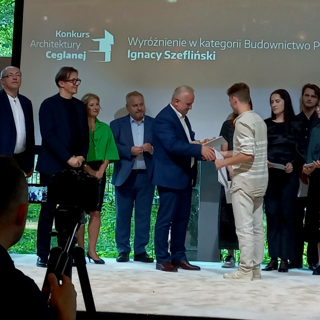 Na pierwszym planie mężczyzna w jasnym ubraniu odbiera dyplom od elegancko ubranego mężczyzny w garniturze. W tle stoi kilkanaście osób – kobiety i mężczyźni. W tle na ekranie widnieje napis: „Konkurs Architektury Ceglanej. Wyróżnienie w kategorii Budownictwo Przyszłości – Ignacy Szefliński”. 