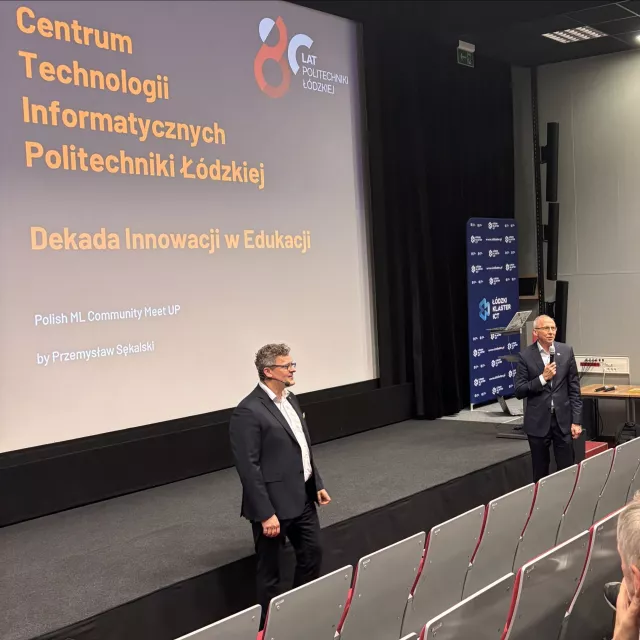 Dwóch mężczyzn stoi przed publicznością siedzącą na widowni, jeden z nich przemawia do mikrofonu