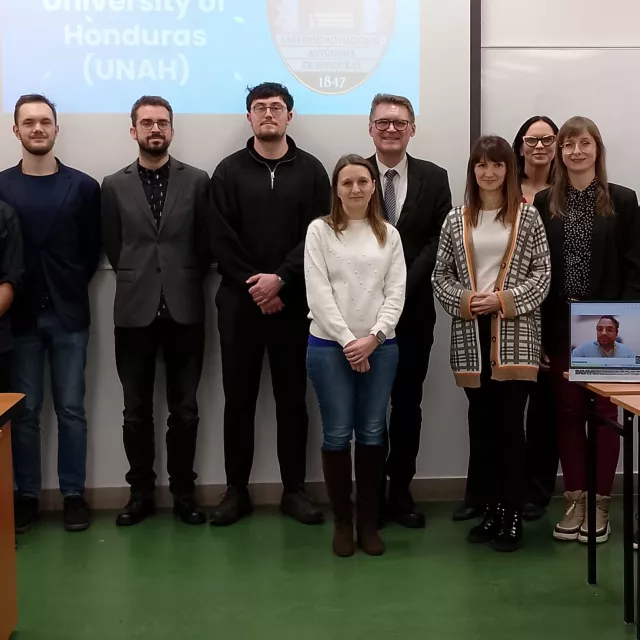 Kilkanaście osób stoi na tle ściany w sali wykładowej, za nimi wyświetla się slajd prezentacji