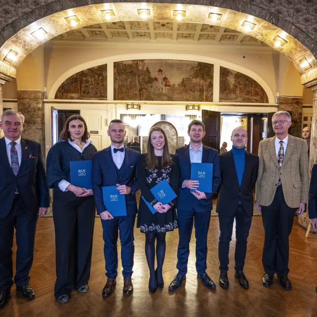 Grupa ośmiu osób stoi w zabytkowym wnętrzu, kilkoro z nich trzyma w ręku dyplomy