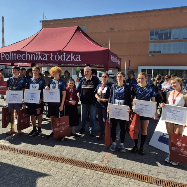 Młodzi ludzie w sportowych podkoszulkach z dyplomami w ręku i medalami na szyi, w towarzystwie małżonki profesora i rektora. Z tyłu stoi namiot z napisem Politechnika Łódzka i budynek uczelni 